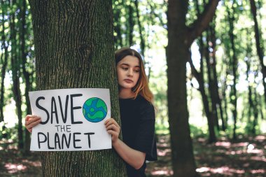 Genç bir kadın, ormandaki gezegeni, doğayı, temizliği, geri dönüşümü ve ekolojiyi kurtarma çağrısını içeren bir poster tutuyor..