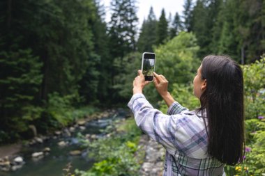 Genç bir kadın ormanın dağlarında akıllı bir telefonla fotoğraf çekiyor. Vahşi doğada seyahat kavramı..