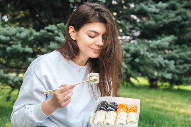 Cheerful young woman eating sushi in the park, picnic in nature, sushi delivery concept.