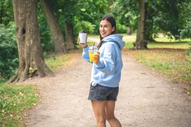 Kapüşonlu, şortlu, portakal suyu ve bir fincan kahveyle parkta yürüyen genç bir kadın..