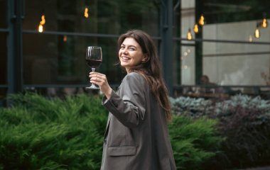 Attractive young woman with glass of red wine outside near restaurant on blurred background, party and celebration concept.