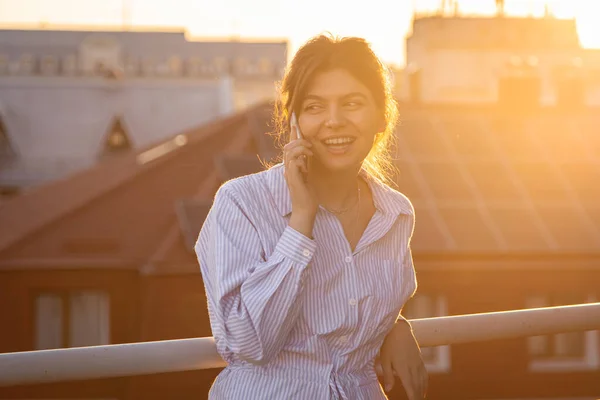 A young woman is talking on the phone on the balcony at sunset on a hot summer evening.