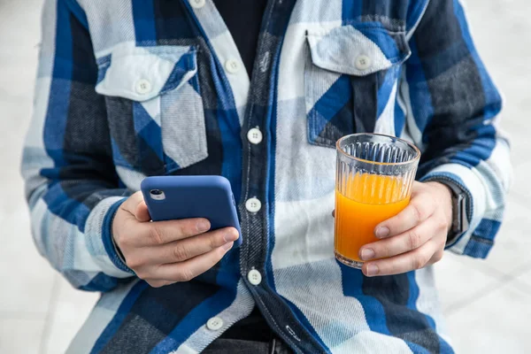 A man in a blue plaid shirt uses a smartphone and drinks orange juice.