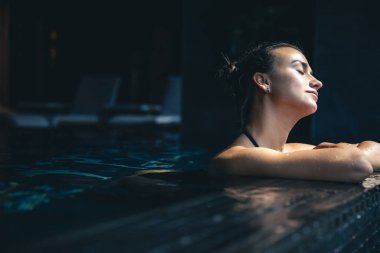 Attractive young woman in a black swimsuit is relaxing in the pool in the spa complex, copy space.