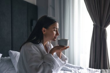 A young woman in a white bathrobe with a cup of coffee in bed in the morning.