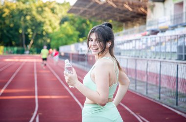 Spor kıyafetli çekici genç bir kadın stadyumda bir şişe suyla koşuyor..