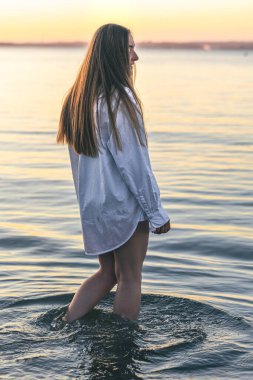 A woman in a bathing suit and a white shirt in the sea at sunset, summer concept.