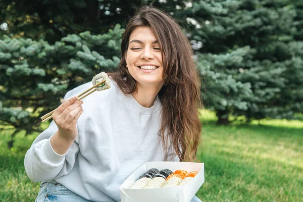 Cheerful young woman eating sushi in the park, picnic in nature, sushi delivery concept.
