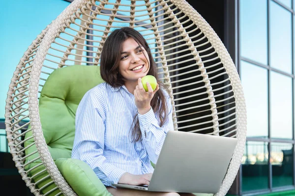 A young woman eats an apple and works on a laptop while sitting in a hammock on the office terrace.