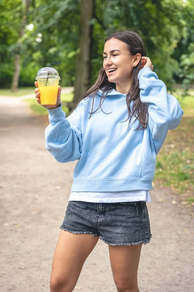 A young woman in hoodie with shorts on a walk in the park with orange juice.