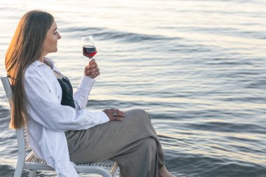 A beautiful woman with a glass of wine is resting on the seashore, sitting on a chair at sunset.
