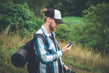 A male traveler with a backpack, a mat and a water bottle looks into a smartphone.