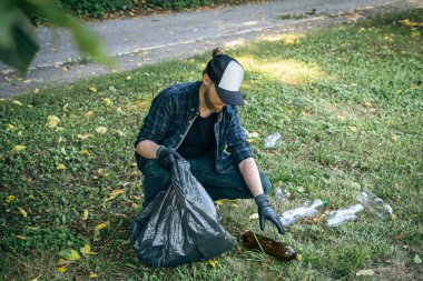 A young man with a garbage bag in the forest cleans up plastic bottles, the concept of love for nature and care for the ecology.