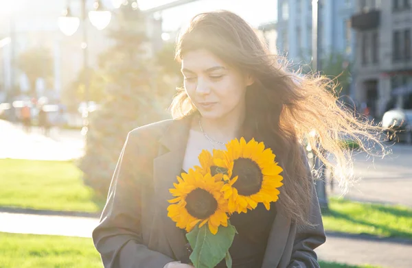 A young woman with a bouquet of sunflowers in the sun at sunset in the city.