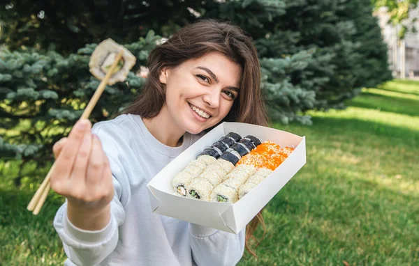 Cheerful young woman eating sushi in the park, picnic in nature, sushi delivery concept.