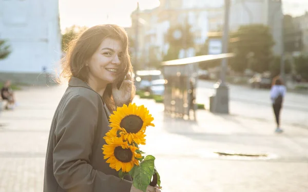 A young business woman with a bouquet of sunflowers talking on the phone at sunset in the city.