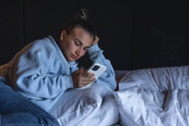 A young tired woman uses smartphone while lying in bed in the evening.