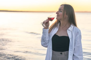 Beautiful woman with a glass of wine against the backdrop of the sea at sunset, copy space.