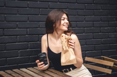 A young woman with a croissant and a cup of coffee on the background of a black brick wall of the cafe exterior.