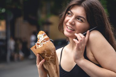 A young woman with a croissant and a cup of coffee on the background of a black brick wall of the cafe exterior.