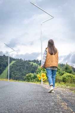 Elinde bir buket ayçiçeği olan genç bir kadın dağlarda asfalt bir yolda yürüyor..