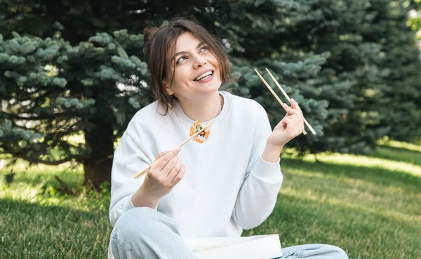 Cheerful young woman eating sushi in the park, picnic in nature, sushi delivery concept.