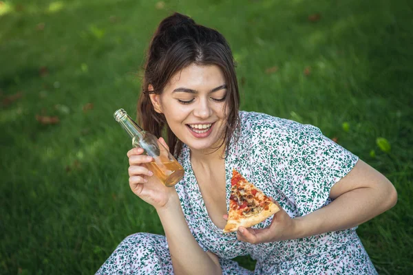 Portrait of a young woman with pizza and a bottle of drink in a picnic on the grass.