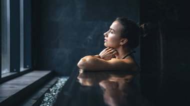 Attractive young woman in a black swimsuit is relaxing in the pool in the spa complex, copy space.