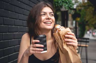 A young woman with a croissant and a cup of coffee on the background of a black brick wall of the cafe exterior.