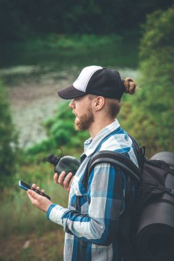A male traveler with a backpack, a mat and a water bottle looks into a smartphone.