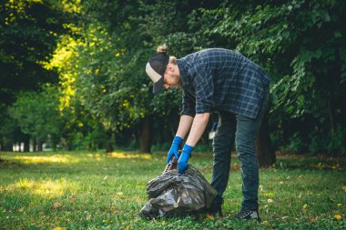 A young man with a garbage bag in the forest cleans up plastic bottles, the concept of love for nature and care for the ecology, copy space.