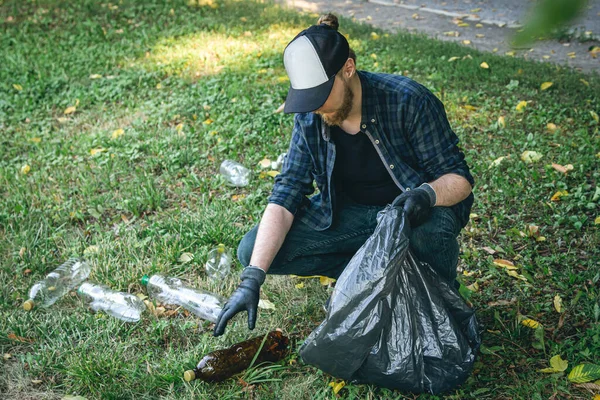 A young man with a garbage bag in the forest cleans up plastic bottles, the concept of love for nature and care for the ecology.
