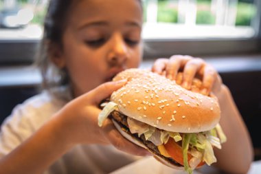 A little girl eats a big appetizing burger, close-up.