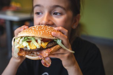 A little girl eats a big appetizing burger in a cafe, close-up.