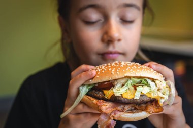 A little girl eats a big appetizing burger in a cafe, close-up.