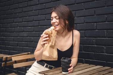 A young woman with a croissant and a cup of coffee on the background of a black brick wall of the cafe exterior.