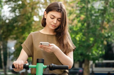 Attractive young woman with a smartphone in her hands rents an electric scooter on a sunny day.
