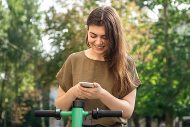 Attractive young woman with a smartphone in her hands rents an electric scooter on a sunny day.