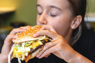 A little girl eats a big appetizing burger in a cafe, close-up.
