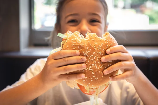 A little girl eats a big appetizing burger, close-up.