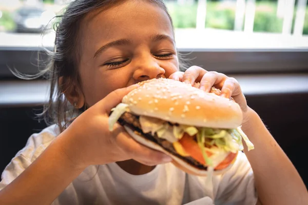 A little girl eats a big appetizing burger, close-up.