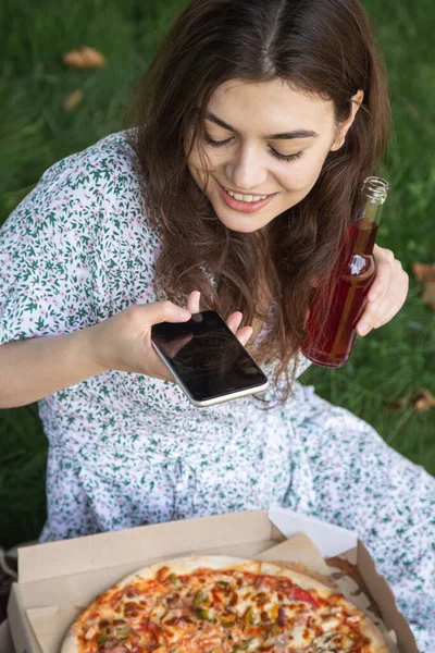 A young woman taking a photo of pizza in nature, picnic concept.