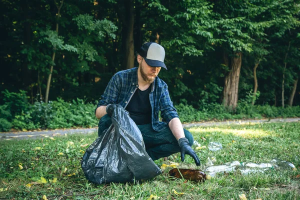 A young man with a garbage bag in the forest cleans up plastic bottles, the concept of love for nature and care for the ecology.