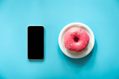 Pembe kremalı çörek ve mavi arka planda akıllı telefon izole edilmiş, kavramsal minimalizm..