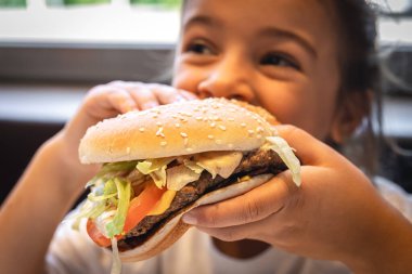 A little girl eats a big appetizing burger, close-up.