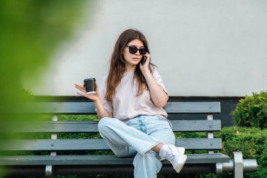 Business young woman sits on a bench with a cup of coffee and speaks on the phone.