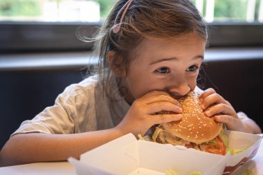 A little girl eats a big appetizing burger in a cafe, close-up.