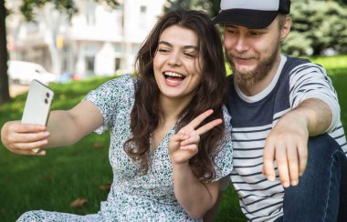 Happy boyfriend and girlfriend take a selfie while sitting on the grass in the park.