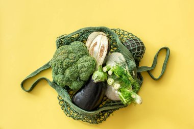 Fresh green vegetables in a green string bag on a yellow background, flat lay.