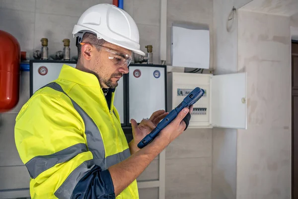 Man, an electrical technician in uniform working in a switchboard with fuses. Installation and connection of electrical equipment. Professional uses a tablet.